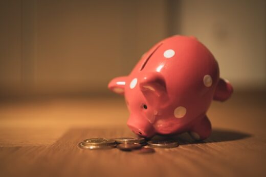 Pink piggy bank leaning toward coins on a wooden floor, symbolizing saving money through Health Savings Accounts (HSAs).